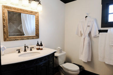 Elegant bathroom with dark carved vanity and marble countertop sink, ornate gold-framed mirror, wall sconces, hanging white bathrobe and towels next to a toilet