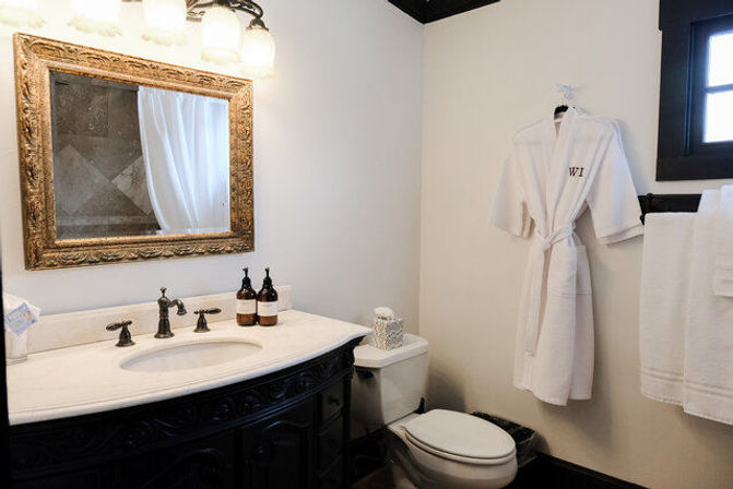 Elegant bathroom with dark carved vanity and marble countertop sink, ornate gold-framed mirror, wall sconces, hanging white bathrobe and towels next to a toilet