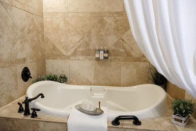Spa-style bathroom with beige stone tiles and a white soaking tub, oil-rubbed bronze faucet and handles, white waffle shower curtain, towel with soap tray, and potted greenery