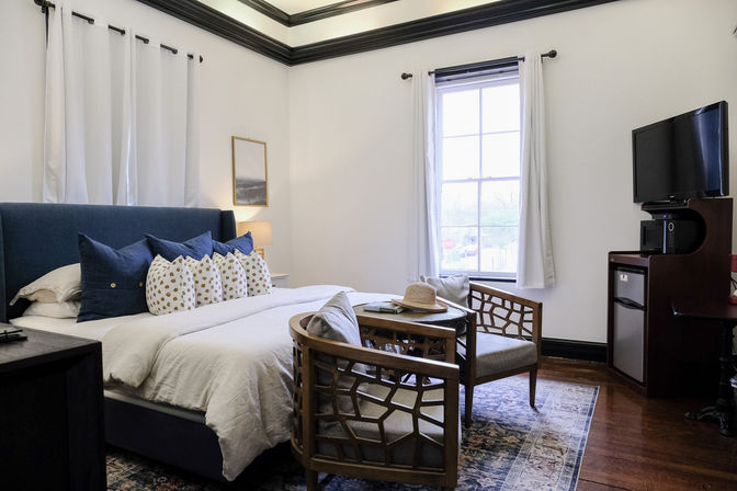 Cozy modern guest bedroom with navy upholstered headboard, white bedding and patterned pillows, wooden lattice chairs around a small table with a straw hat, sunlit window with white curtains, wall-mounted TV and mini-fridge on hardwood floors.