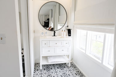 Bright white bathroom with a vintage-style vanity topped by a round black-framed mirror, patterned black-and-white tile floor, soap dispensers and folded towel, and a sunlit window with a white Roman shade.