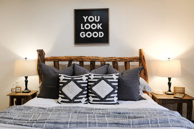 Cozy bedroom interior with rustic log headboard, patterned black-and-white throw pillows, gray bedding, matching wooden bedside tables and lamps, and framed wall art that reads "YOU LOOK GOOD".