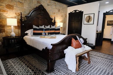 Ornate carved dark-wood bed in a cozy boutique hotel bedroom with exposed stone accent wall, patterned rug, bedside lamps, striped pillows, and a bench draped with a throw and hat.