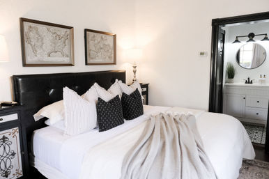 Cozy modern guest bedroom with black tufted headboard, layered white bedding, striped and patterned accent pillows, soft gray throw, framed vintage maps above the bed, matching nightstands with lamps, and a visible en-suite bathroom with round mirror and vanity.