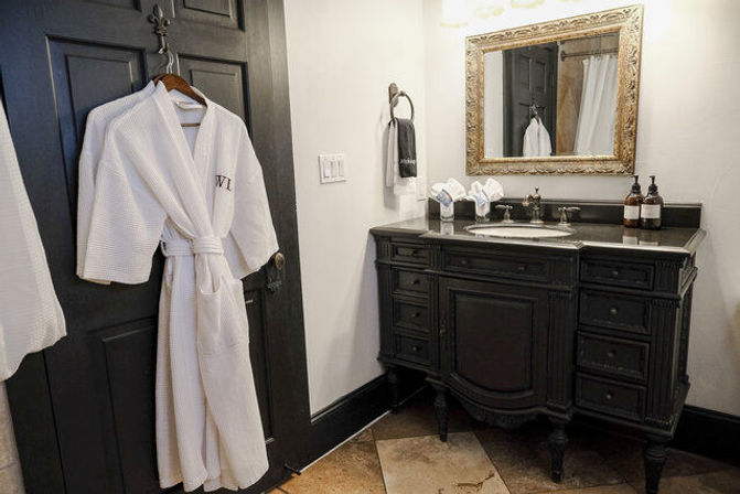 Spa-style bathroom with a white waffle robe hanging on a black paneled door, ornate gold-framed mirror above a dark vintage vanity with sink, amber soap dispensers and tiled floor.