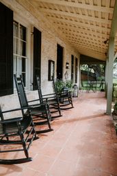 Covered stone farmhouse porch with terracotta tile floor, three black wooden rocking chairs by shuttered windows, lanterns, potted greenery and an iron bistro table overlooking the yard.