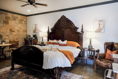 Ornate dark-wood king bed in a cozy rustic bedroom with stone accent wall, warm orange bedding, leather armchair, bedside lamps, ceiling fan and a decorative tray on the bed.