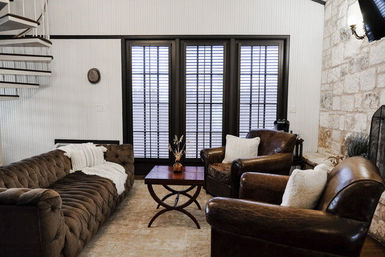 Cozy modern farmhouse living room with a tufted brown sofa, two leather armchairs, wooden coffee table, tall shuttered windows, stone fireplace, and a spiral staircase.