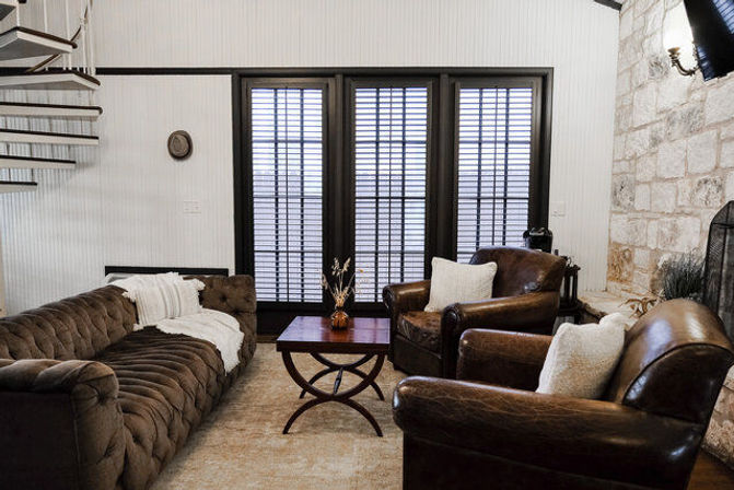 Cozy modern farmhouse living room with a tufted brown sofa, two leather armchairs, wooden coffee table, tall shuttered windows, stone fireplace, and a spiral staircase.