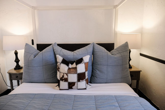 Cozy bedroom with blue quilted pillows and comforter, a cowhide patchwork accent pillow centered on a black headboard, wooden nightstands and lit table lamps