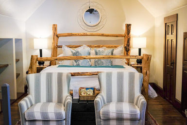 Cozy rustic cabin bedroom with a log-framed bed, tropical-print throw pillows, round wall mirror, two bedside lamps, striped upholstered armchairs at the foot of the bed and hardwood floors
