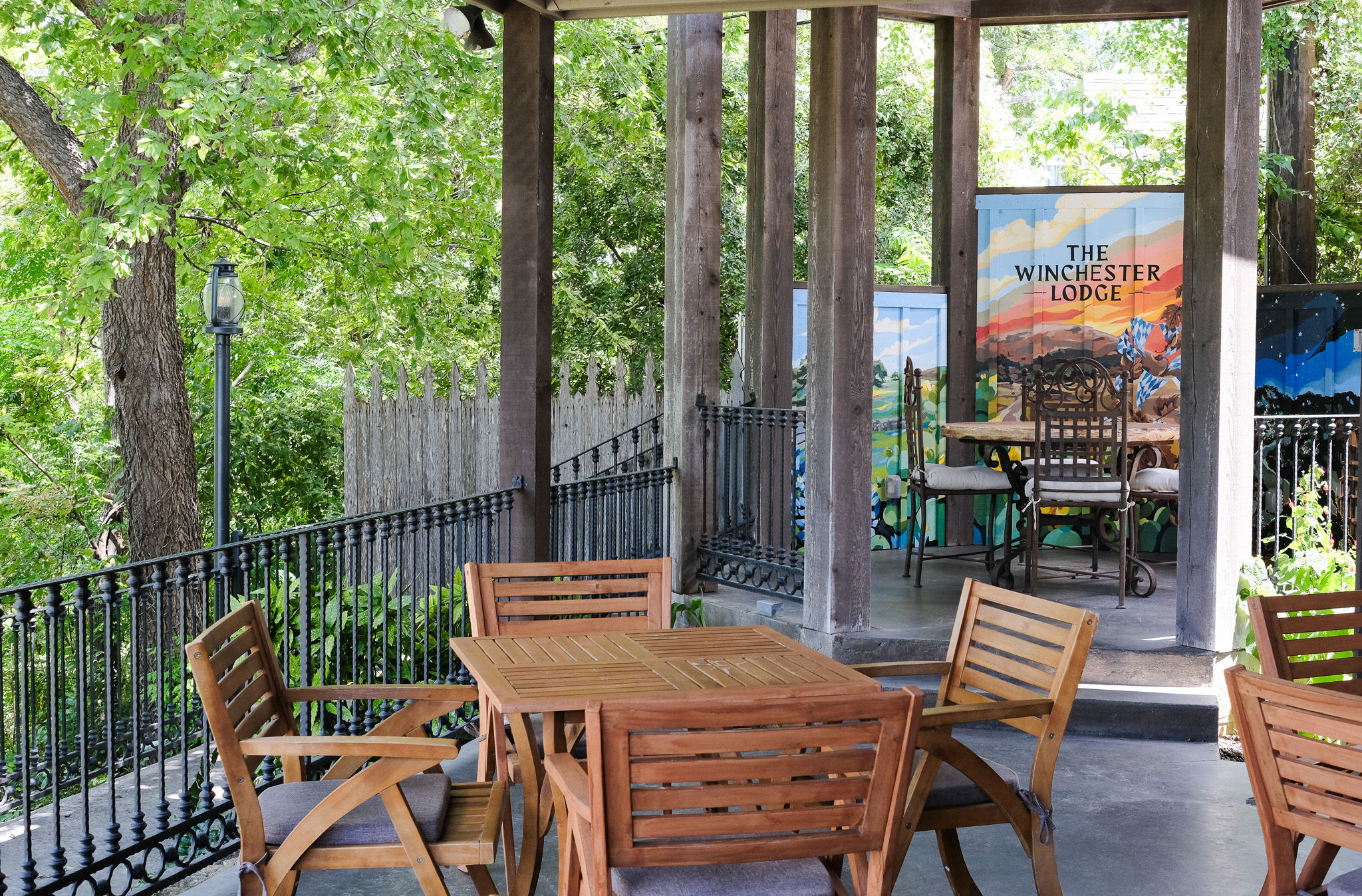 Cozy shaded outdoor patio with a wooden dining set, wrought-iron railing, colorful painted privacy panels, and lush green trees surrounding a terrace.