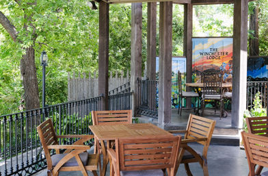 Cozy shaded outdoor patio with a wooden dining set, wrought-iron railing, colorful painted privacy panels, and lush green trees surrounding a terrace.