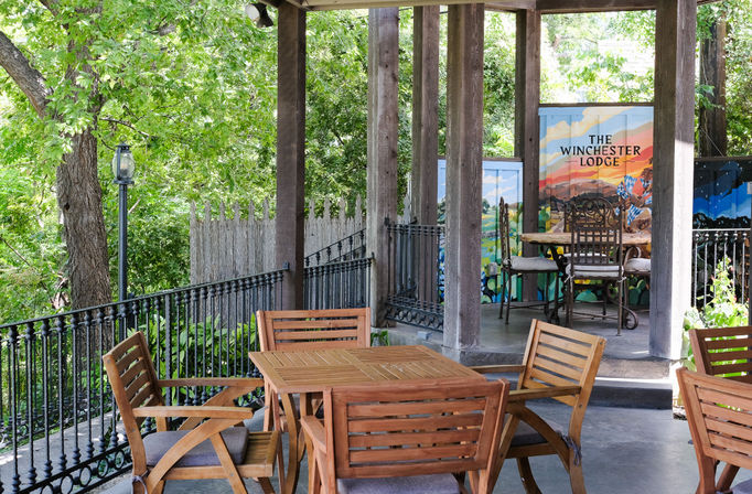 Cozy shaded outdoor patio with a wooden dining set, wrought-iron railing, colorful painted privacy panels, and lush green trees surrounding a terrace.