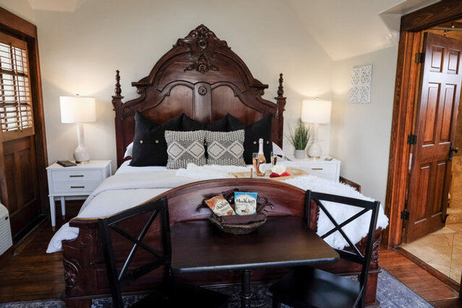 Cozy guestroom with an ornate carved dark-wood bed, white linens and patterned pillows, matching nightstands with lamps, a small table with two chairs and a snack basket, and a champagne tray on the bed.