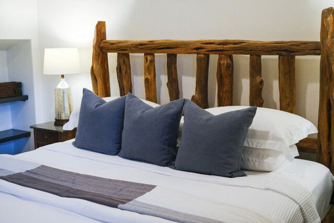 Cozy bedroom with rustic log headboard, crisp white linens, three navy throw pillows and a bedside lamp on a wooden nightstand.