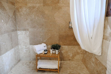 Spa-like beige stone shower with a wooden bench holding rolled white towels, toiletries and a small potted plant, and a white shower curtain.