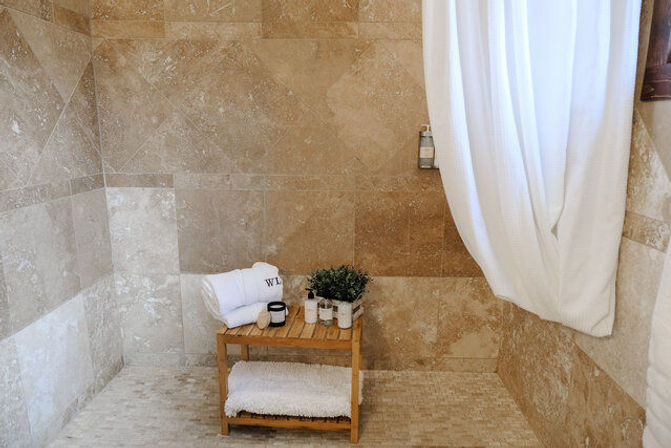 Spa-like beige stone shower with a wooden bench holding rolled white towels, toiletries and a small potted plant, and a white shower curtain.