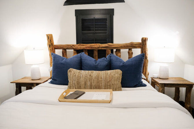 Cozy rustic bedroom interior with natural log headboard, white bedding, three navy throw pillows, woven lumbar cushion, wooden nightstands with white lamps, and a tray on the bed.