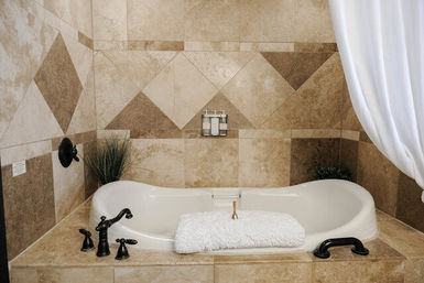 Inviting spa-style master bathroom alcove with white soaking tub set into beige stone tile surround, oil-rubbed bronze faucets, folded fluffy white towel and potted greenery.