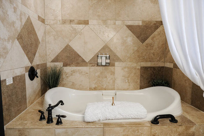 Inviting spa-style master bathroom alcove with white soaking tub set into beige stone tile surround, oil-rubbed bronze faucets, folded fluffy white towel and potted greenery.