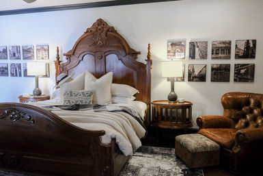 Cozy boutique bedroom with ornate carved wooden bed, soft neutral bedding and patterned pillows, bedside lamps, gallery wall of black-and-white photos, and a tufted leather armchair with ottoman.