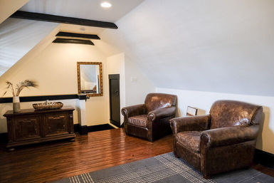Cozy attic sitting room with two brown leather club chairs, hardwood floors, antique wooden chest, framed mirror and sloped white ceiling with exposed black beams.