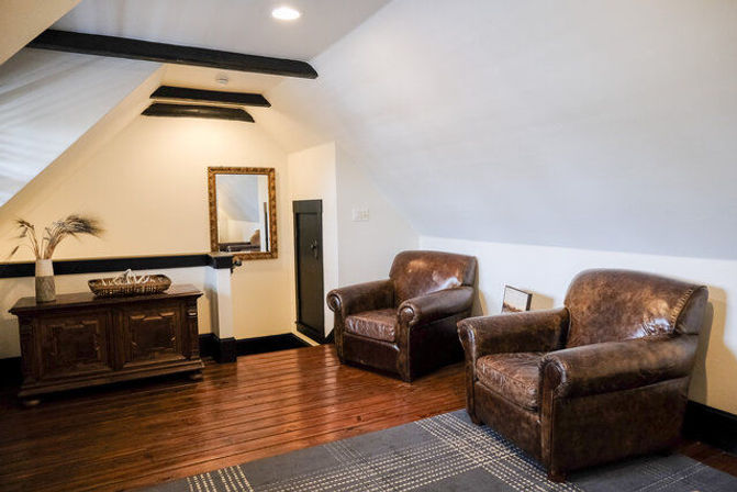 Cozy attic sitting room with two brown leather club chairs, hardwood floors, antique wooden chest, framed mirror and sloped white ceiling with exposed black beams.