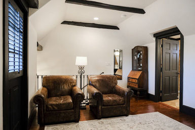 Cozy upstairs sitting area with two worn brown leather armchairs, white sloped ceiling with dark exposed beams, wood floors, standing lamp, vintage wooden secretary cabinet and light area rug.