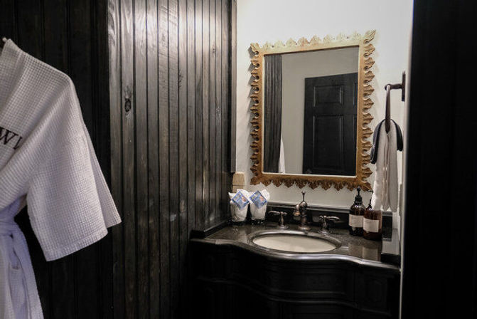 Moody dark wood–paneled bathroom with black vanity and oval sink, ornate gold-framed mirror, white waffle robe hanging and amber soap bottles on the counter