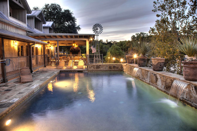 Twilight-lit outdoor pool with stone spillways and potted yuccas beside a cozy rustic farmhouse porch glowing with hanging lanterns, lounge chairs and a windmill in the wooded background.