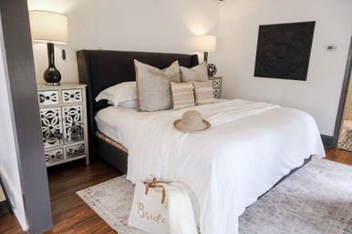 Cozy, light-filled neutral bedroom with crisp white linens and layered pillows, straw sun hat on the bed, mirrored nightstands with lamps, and a 'Bride' canvas tote on the floor.