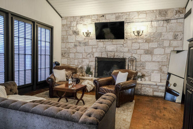 Cozy rustic-chic living room with exposed stone fireplace and wall-mounted TV, tufted gray sofa, two brown leather armchairs around a wooden coffee table, shuttered glass doors and warm wood floor.
