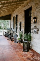 Inviting covered rustic stone farmhouse porch with terracotta tile floor, a row of black wooden rocking chairs, large lanterns and potted plants flanking a dark front door and decorative house-number plaque.