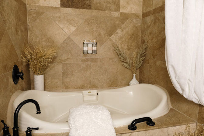 Spa-style beige tiled corner bathtub with black faucets, white towel draped over the rim, dried-grass vases and wall-mounted soap dispensers, white shower curtain
