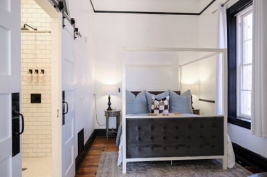 Bright, cozy modern farmhouse-style bedroom with a white four-poster bed, tufted gray footboard, blue pillows, bedside lamps, hardwood floors, black-trim window and a barn door to a subway-tiled shower.