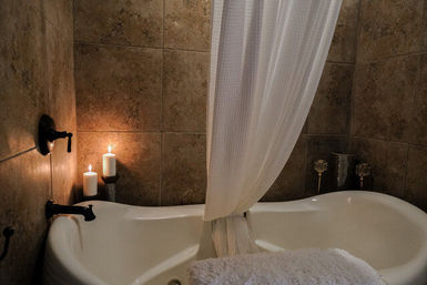Cozy spa-style bathroom with a white freestanding tub, tied white shower curtain, lit pillar candles by brown tiled walls and a fluffy towel on the tub edge.
