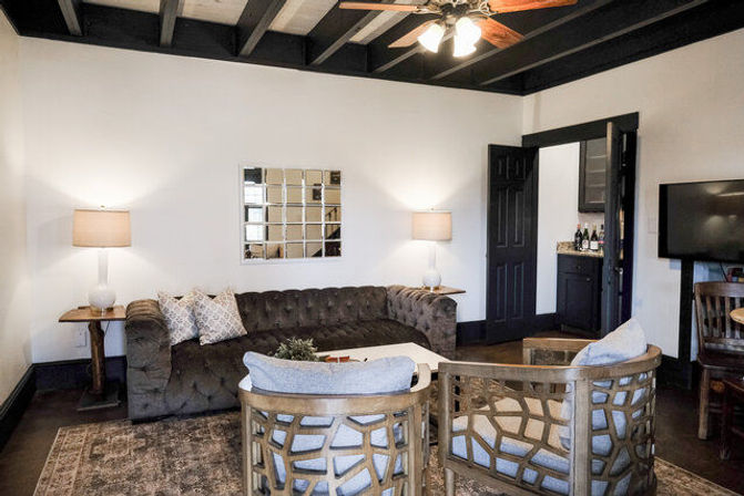 Cozy neutral living room interior with dark exposed ceiling beams and ceiling fan, tufted gray sofa with patterned pillows, mirrored tile accent on a white wall, two wooden lattice-back armchairs, area rug, side tables with lamps and wall-mounted TV.