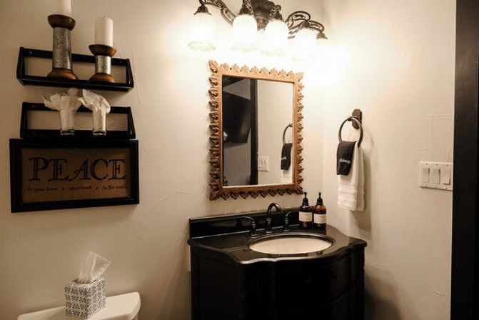 Cozy rustic home bathroom with black vanity and round sink, ornate gold-framed mirror under lit sconces, towel ring, 'PEACE' wall sign, candles and tissue box