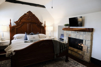 Cozy rustic bedroom with ornate carved wooden bed, white linens, bedside lamps, stone fireplace with wooden mantel and wall-mounted TV
