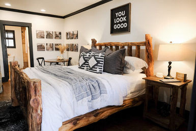 Cozy modern farmhouse bedroom with a rustic live-edge log bed, white linens, patterned throw and pillows, wooden nightstand with lamp, and gallery wall topped by a “You Look Good” sign.