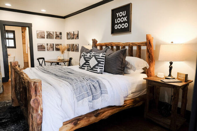 Cozy modern farmhouse bedroom with a rustic live-edge log bed, white linens, patterned throw and pillows, wooden nightstand with lamp, and gallery wall topped by a “You Look Good” sign.