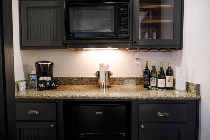 Compact hotel kitchenette with black microwave, single-serve coffee maker and mugs, small mini-fridge, granite countertop, hanging stem glass and assorted wine bottles beneath dark cabinetry.