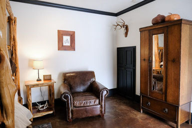 Cozy rustic cabin bedroom corner with brown leather armchair, wooden bedside table and lamp, antler wall mount, and mirrored wooden armoire on a polished floor.
