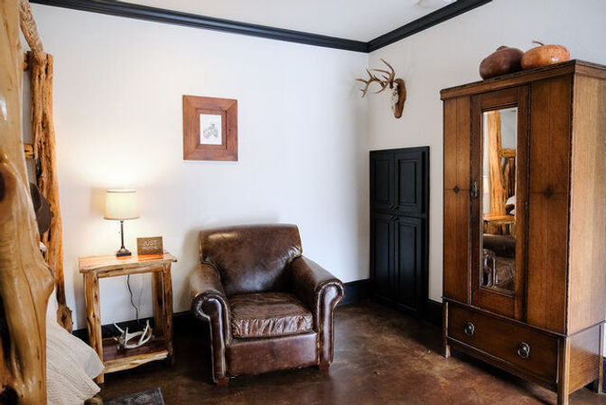 Cozy rustic cabin bedroom corner with brown leather armchair, wooden bedside table and lamp, antler wall mount, and mirrored wooden armoire on a polished floor.