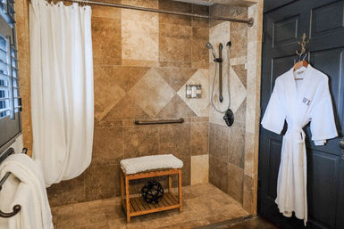 Spa-style walk-in bathroom with warm stone tiles, white shower curtain, wooden bench, handheld showerhead, safety grab bar and white robe hanging on a dark door