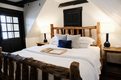 Cozy rustic cabin-style bedroom with a live‑edge log bed, crisp white linens, navy accent pillow, wooden nightstands and lamps beneath a sloped ceiling and shuttered window.