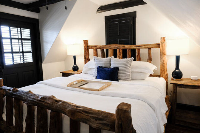 Cozy rustic cabin-style bedroom with a live‑edge log bed, crisp white linens, navy accent pillow, wooden nightstands and lamps beneath a sloped ceiling and shuttered window.