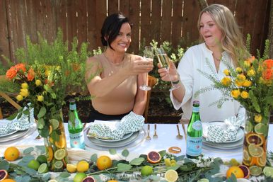 Two friends toasting with champagne flutes at a backyard alfresco brunch table decorated with citrus-filled vases, yellow-and-orange floral arrangements, green patterned napkins, and sparkling water bottles.