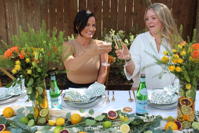 Two friends toasting with champagne flutes at a backyard alfresco brunch table decorated with citrus-filled vases, yellow-and-orange floral arrangements, green patterned napkins, and sparkling water bottles.